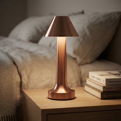 Ladbar bordlampe i rødbronse på nattbord ved siden av en bokstabel i et soverom.