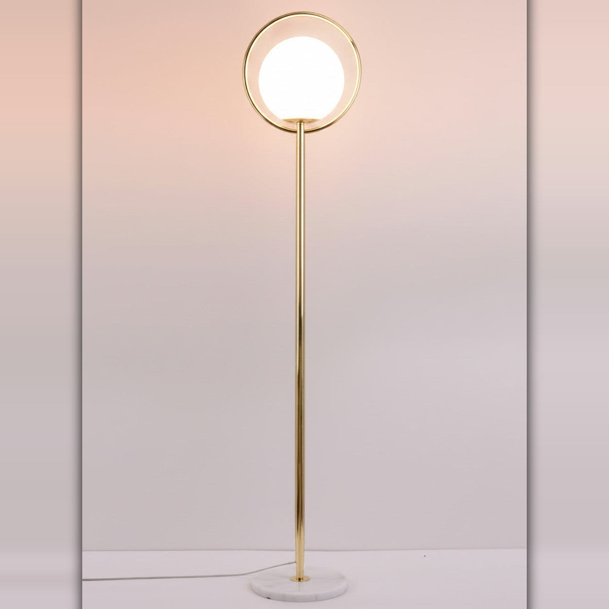 Lysverden gullfarget gulvlampe med sirkulær metallramme og hvit kule i opalglass på marmorfot.