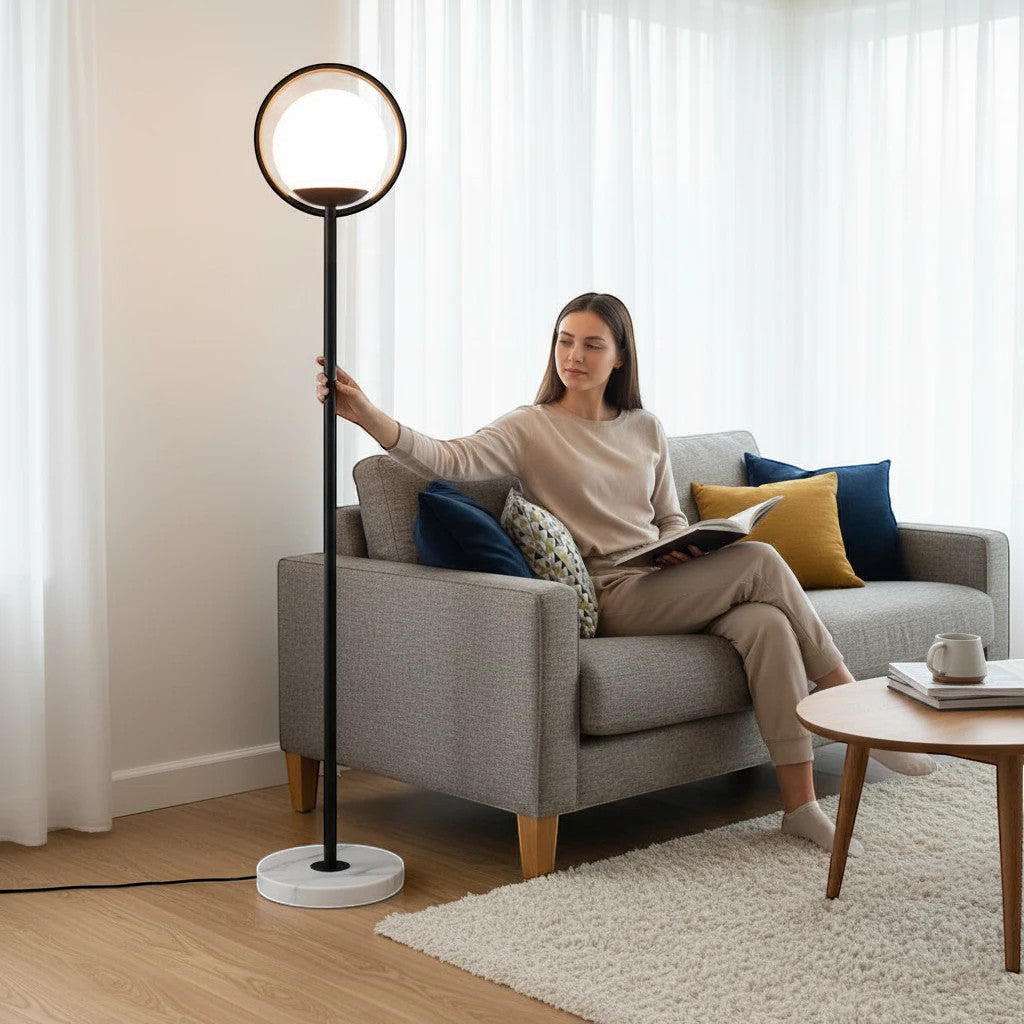 Lysverden sort gulvlampe med opalglasskule og rund metallramme, ved siden av en kvinne i sofaen