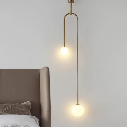 Lysverden minimalistisk pendellampe med to kuler i opalglass på slank messingstang, hengende over en sengegavl.