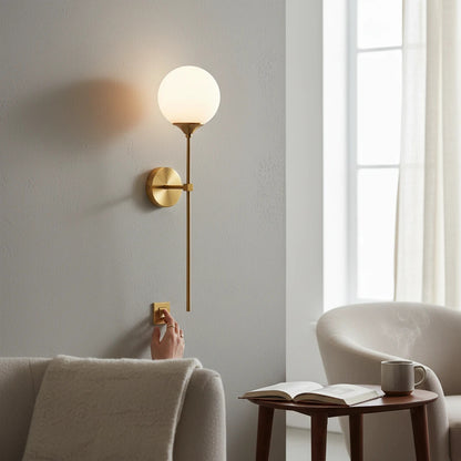Lysverden brass wall lamp with a round opal glass globe shade mounted on a grey wall with a hand turning it on.