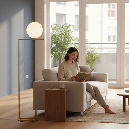 Lysverden gold floor lamp with opal glass globe and integrated side table next to a woman reading on a sofa.