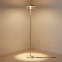 Lysverden elegant floor lamp in gold with a clear glass disk shade casting a warm glow in a minimalist room corner.