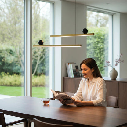 Lysverden linear gold LED pendant light hanging over a wooden dining table with a woman reading a book