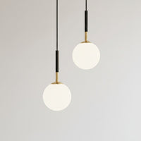 Lysverden minimalistiske pendellamper med melkehvite kuler i opalglass og stammer i sort og gull mot en enkel vegg.