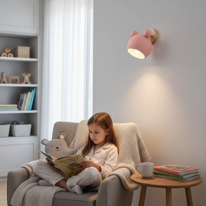Lysverden pink bear-shaped LED wall lamp with wooden detail, mounted in a cozy child's room above a reading chair.