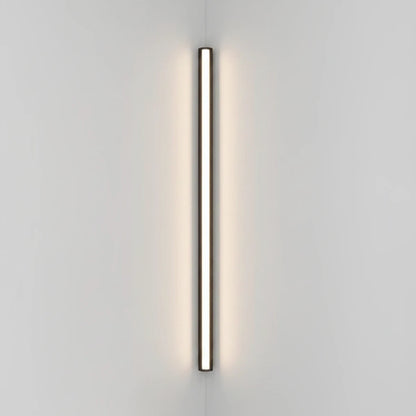 Lysverden minimalistisk sort lineær LED-vegglampe montert vertikalt på en hvit vegg med varmt diffust lys.