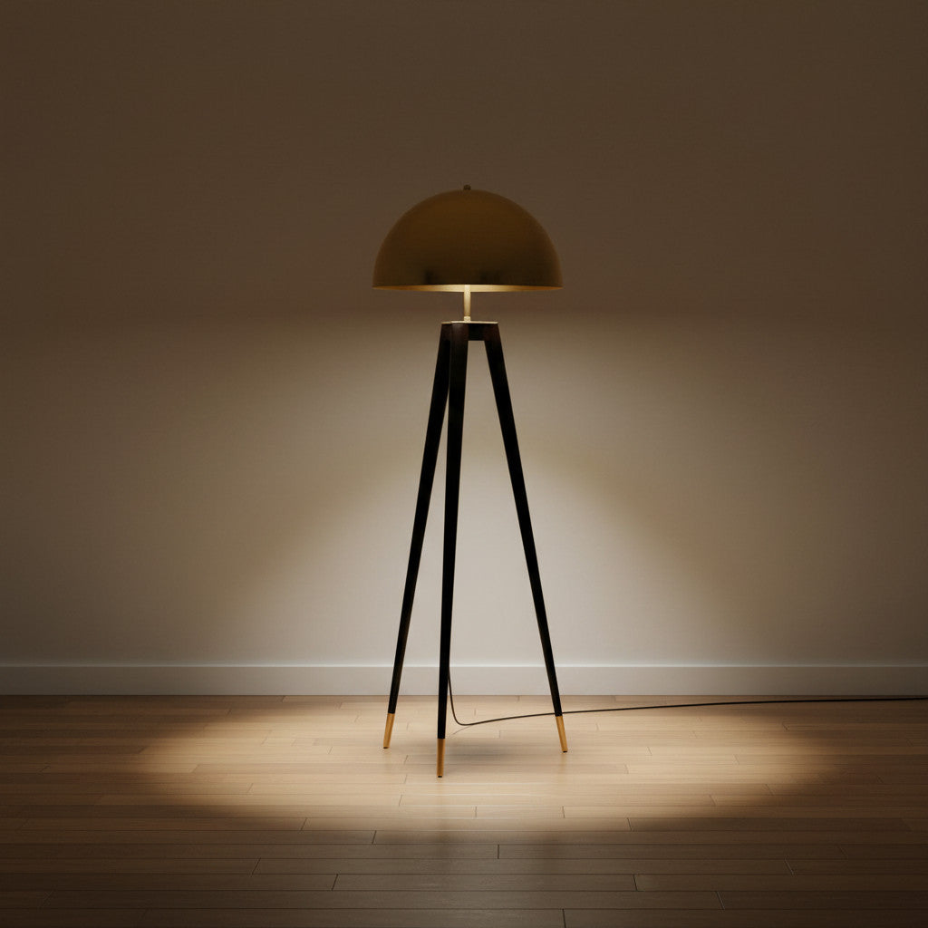 Lysverden tripod floor lamp with a gold brass dome shade and slim black legs casting a warm light on a wooden floor.